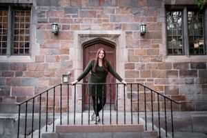 Person in olive green outfit standing on stone steps with metal railings in front of historic building entrance.