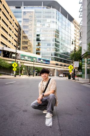 Urban street photo in front of glass building with person crouching in casual attire.
