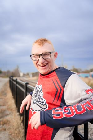 Person wearing red and white varsity jacket smiling while leaning on railing outdoors.