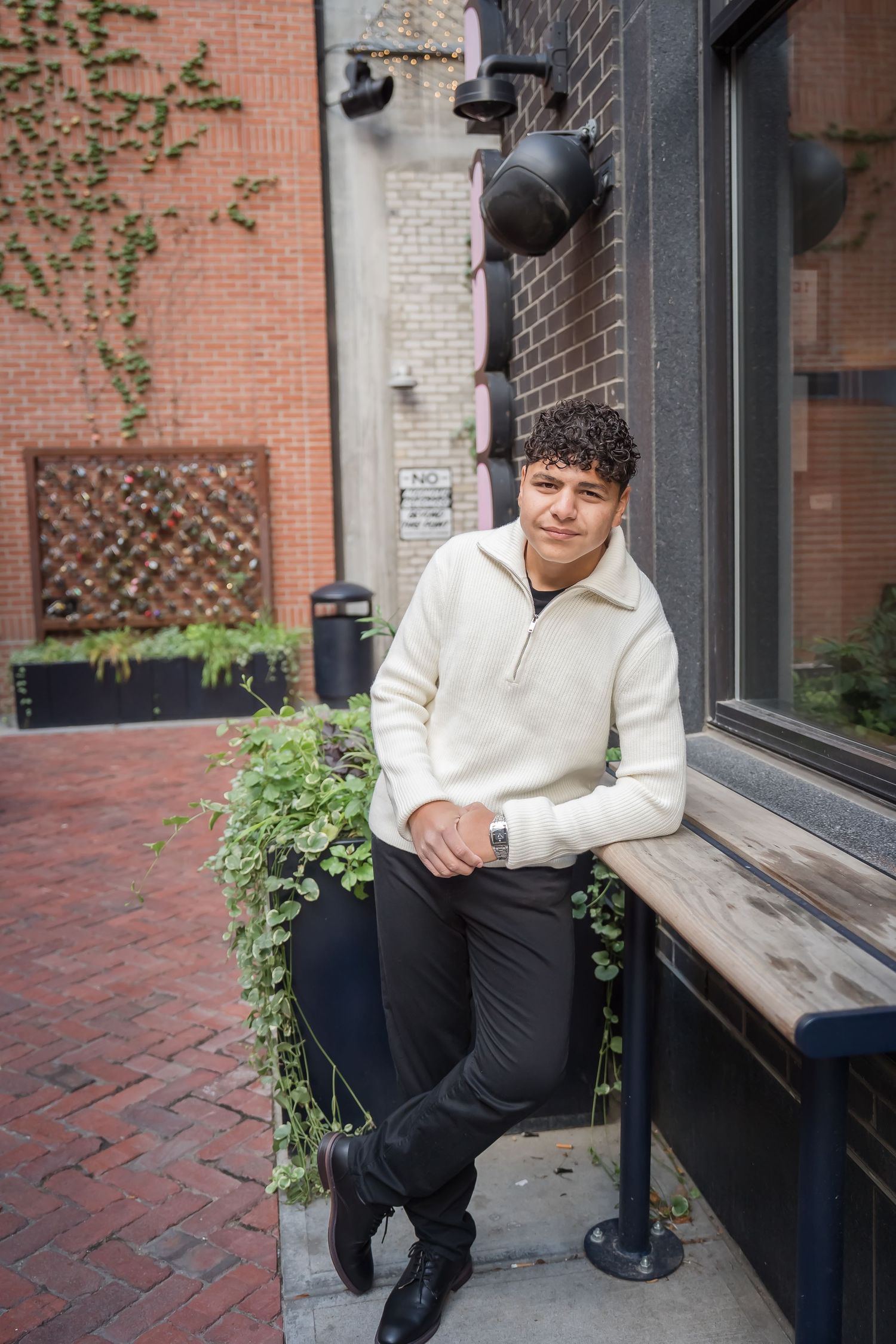 A person in a white sweater sits on a brick ledge outside a building with black window frames and climbing vines.