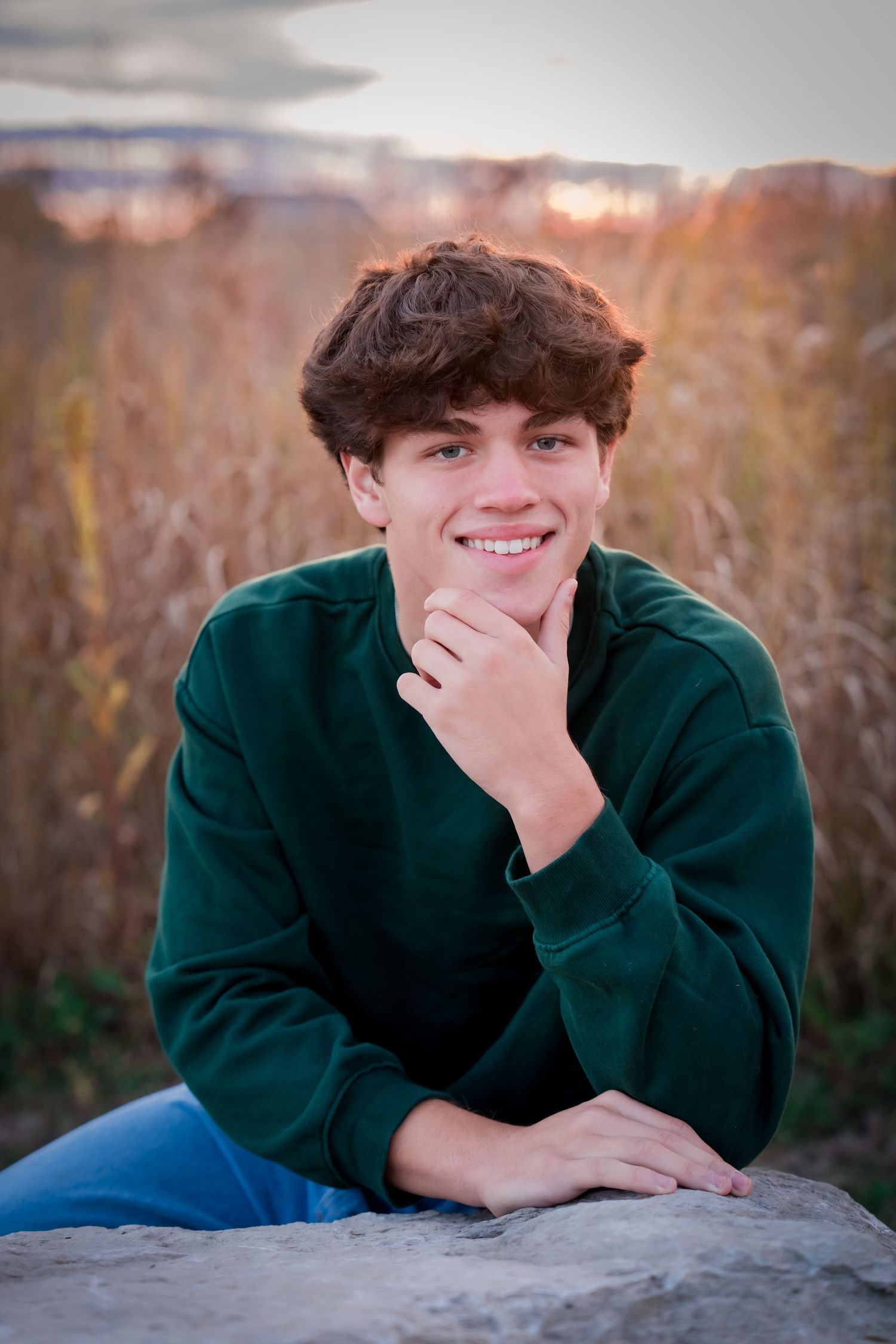 A person in a dark green sweater rests on a rock outdoors during golden hour with autumn foliage in the background.