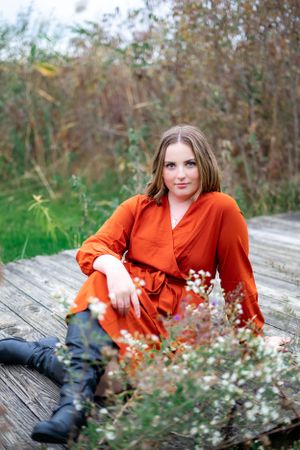 Portrait of someone in a bright orange wrap dress sitting on a wooden deck surrounded by greenery.