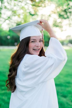 Graduate in white cap and gown poses playfully in a sunny outdoor setting with green grass in the background.