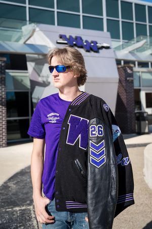 Person wearing purple and black varsity jacket with chevron stripes stands outside a modern building.