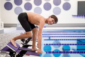 A swimmer performs a butterfly stroke drill in a competition pool with lane lines and timing equipment.