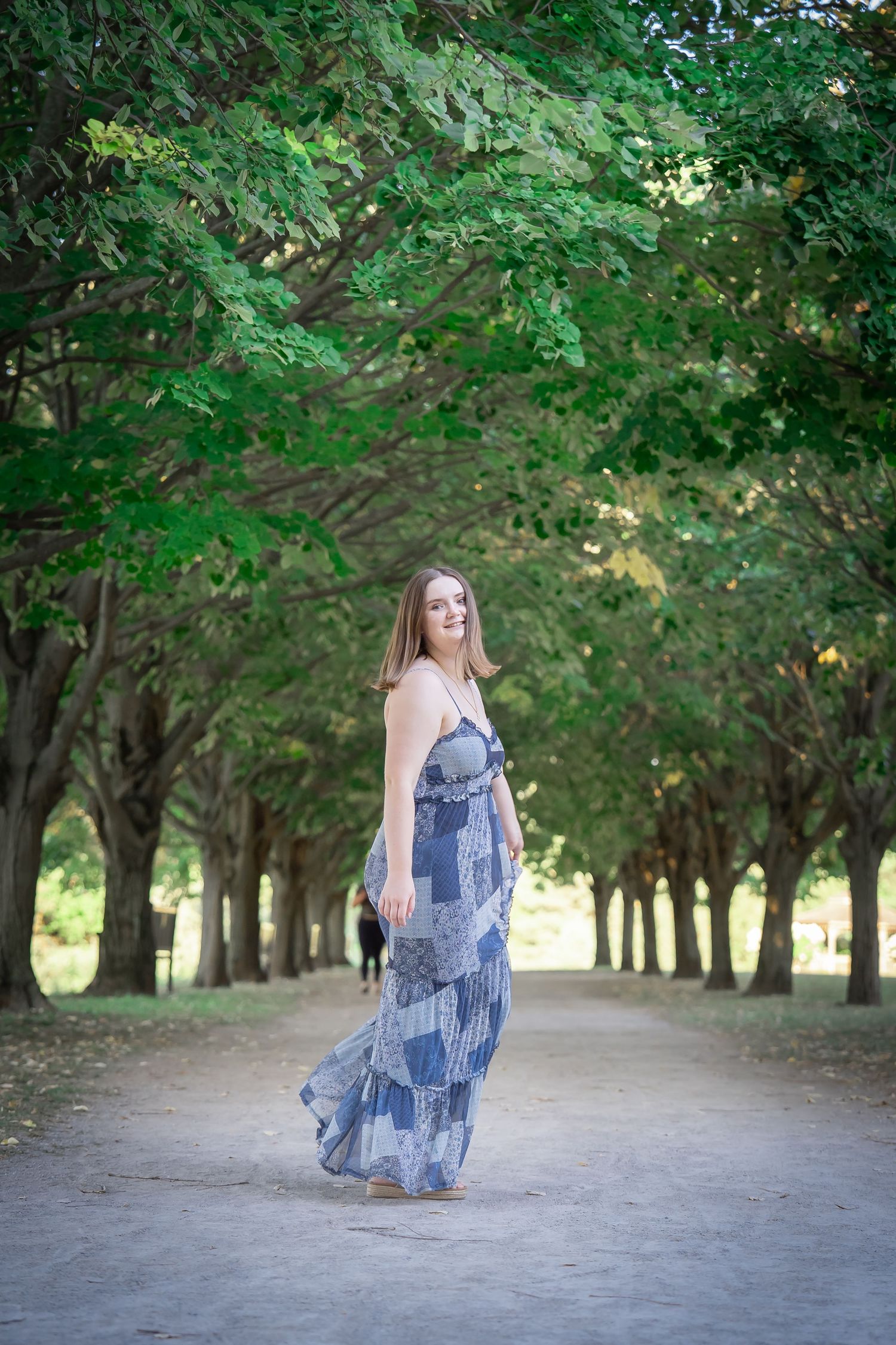 A person in a flowing blue dress poses gracefully in a tree-lined path during a summer photo session.