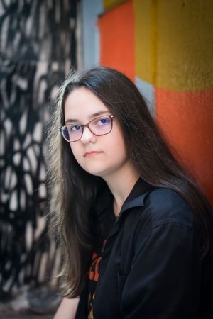 A person with long dark hair and blue glasses gazes upward against a colorful wall background in a creative portrait series.
