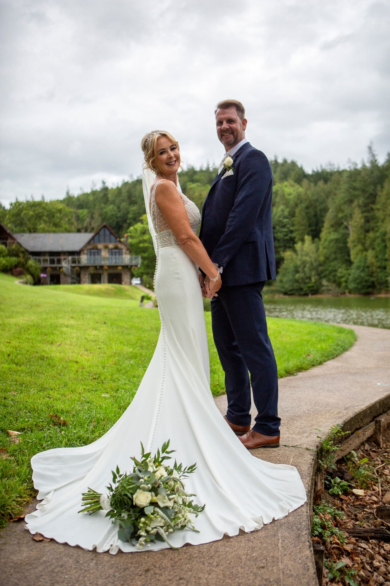 Bride and groom holding hands on a garden path, bride in elegant white gown with train, groom in navy suit.