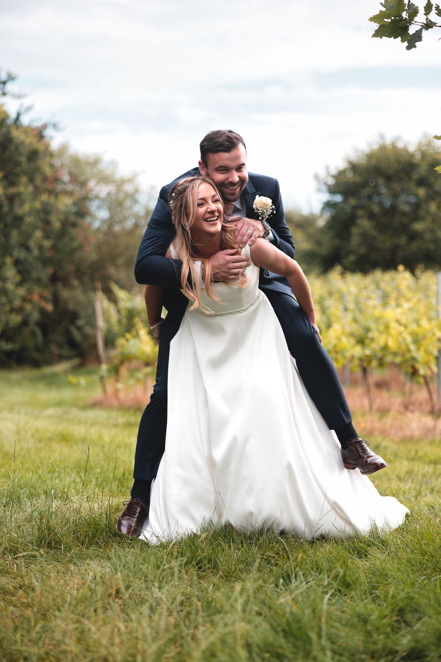 Groom lifting bride playfully on a grass lawn with vineyard countryside backdrop, natural wedding photography Southampton