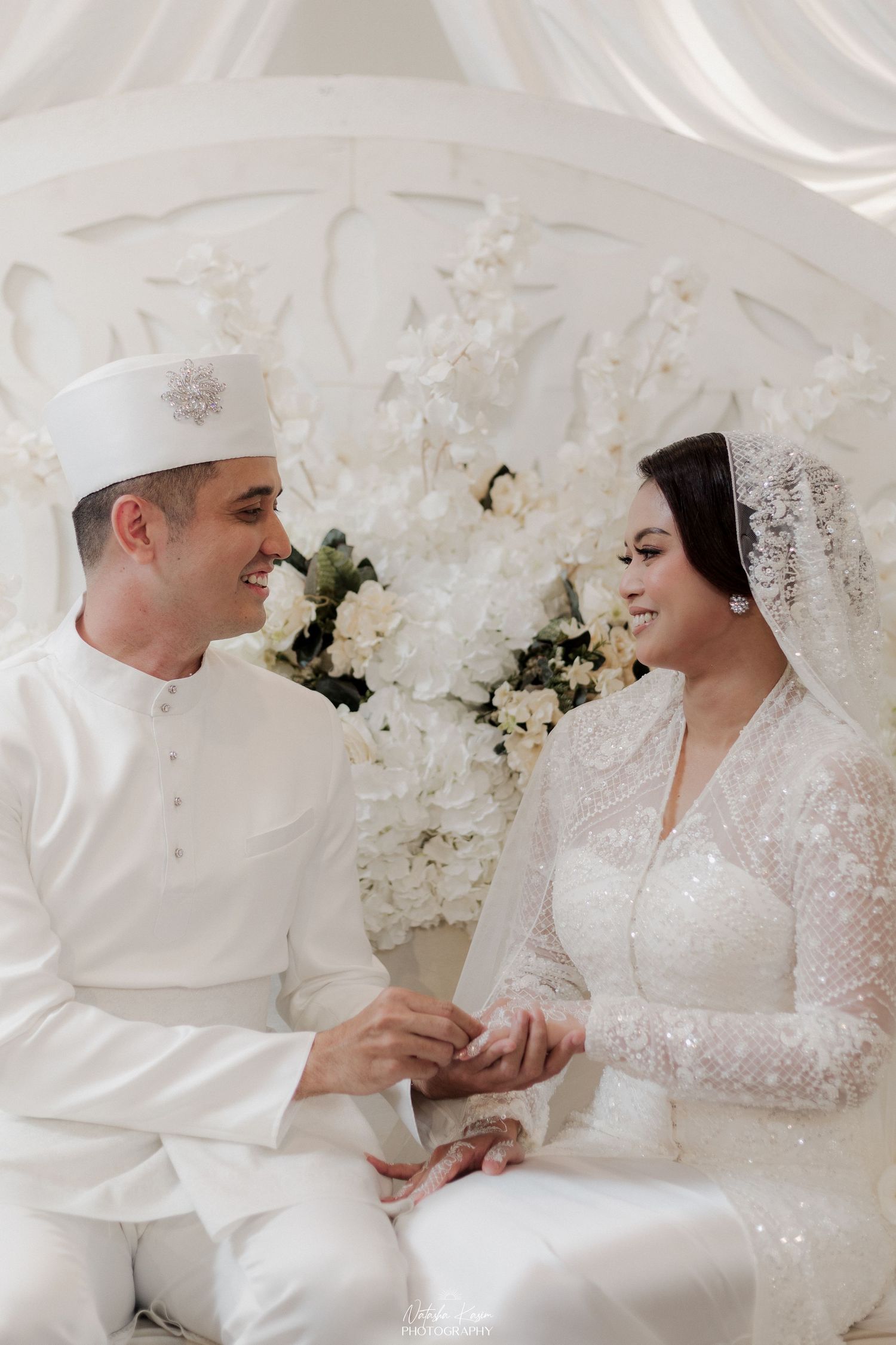 A couple in white traditional wedding attire shares an intimate moment against a floral backdrop.