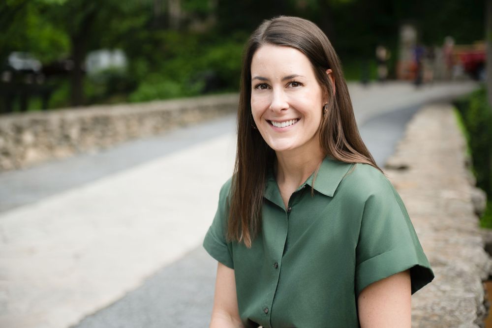 A person in a forest green collared shirt sits outdoors on a curved stone path surrounded by greenery.