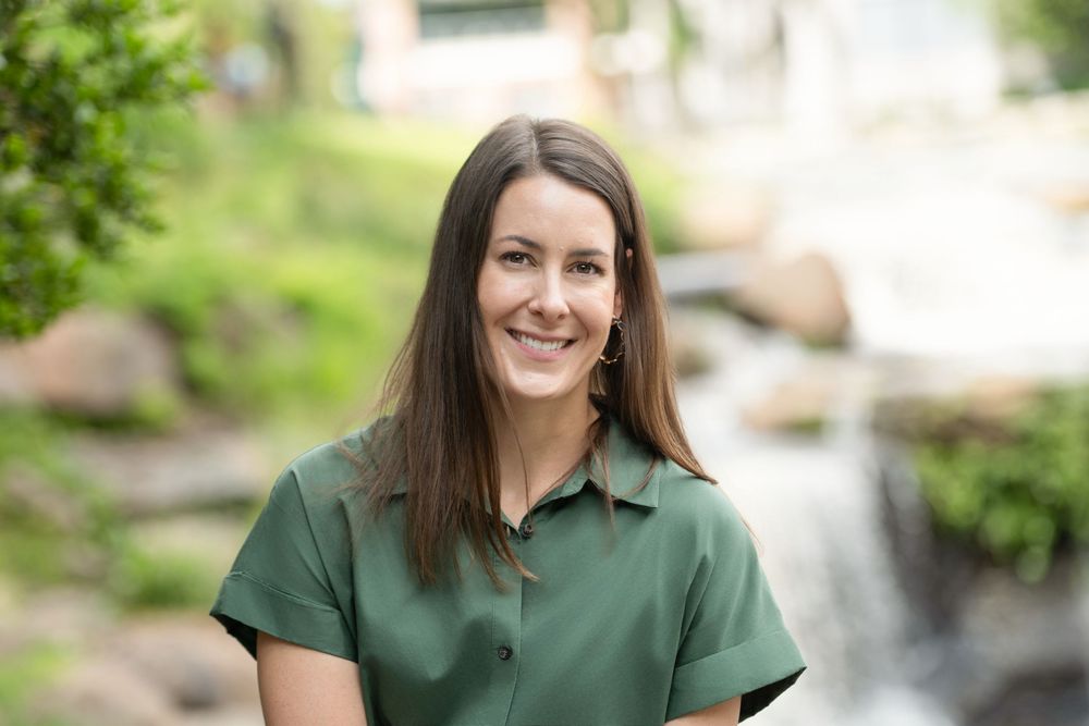 A person in a dark green polo shirt stands smiling outdoors with a waterfall visible in the background.