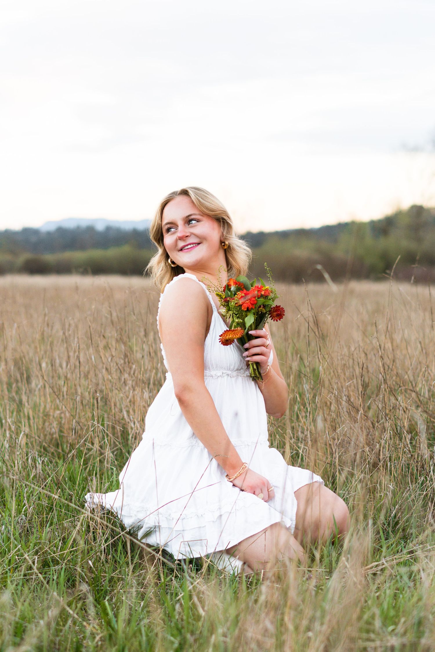 A woman in a white dress sits gracefully in a golden field holding colorful wildflowers at sunset.