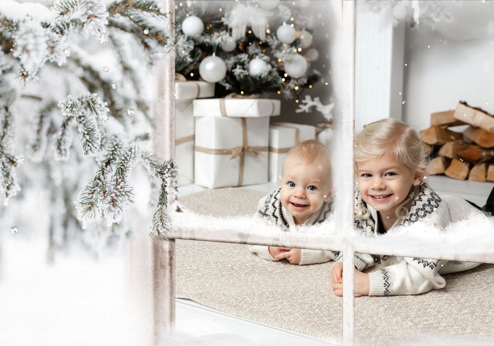Two cheerful toddlers lying by a white window frame with a snowy Christmas tree and wrapped gifts in the background.