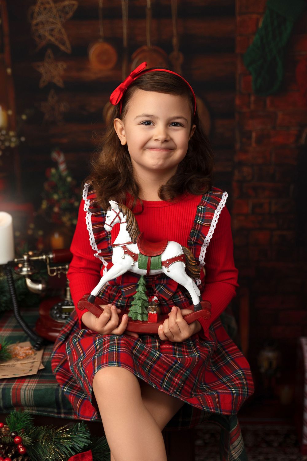 Festive holiday portrait in red dress and plaid skirt with rocking horse decoration.