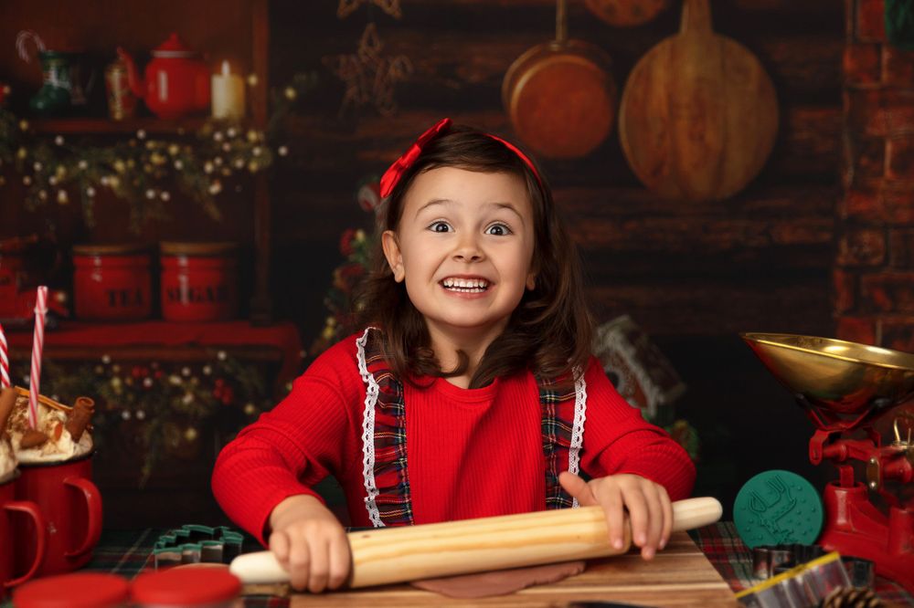 Child in red holiday outfit rolling dough in rustic Christmas kitchen setting.