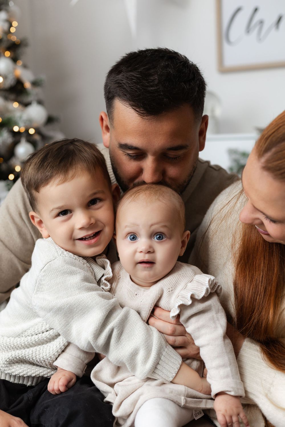 Family with two young children posing near Christmas tree, father kissing baby girl while toddler son smiles.