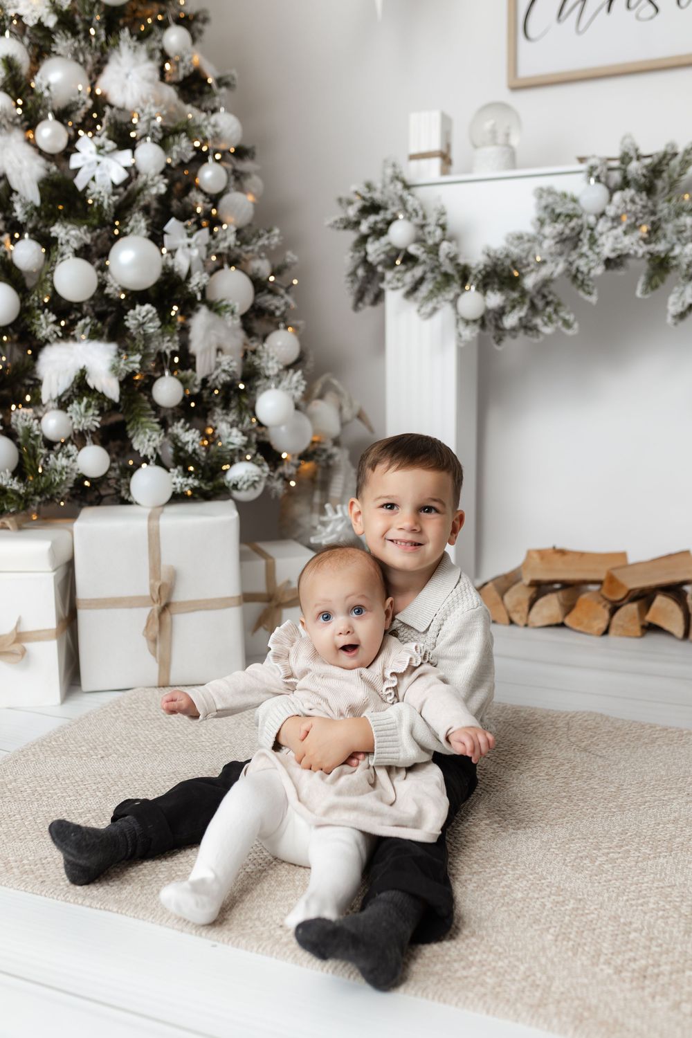 Two young siblings sitting together in front of decorated Christmas tree with white ornaments and gifts.