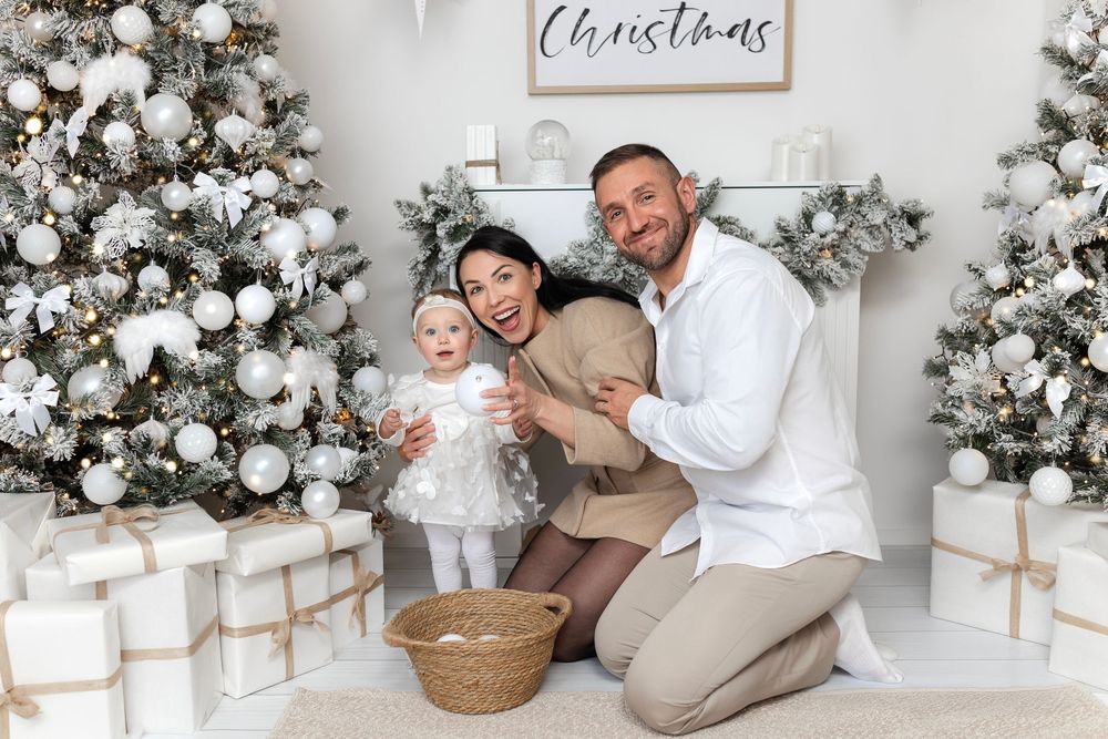 Family Christmas photo with parents and baby girl posing by white decorated trees and gifts.