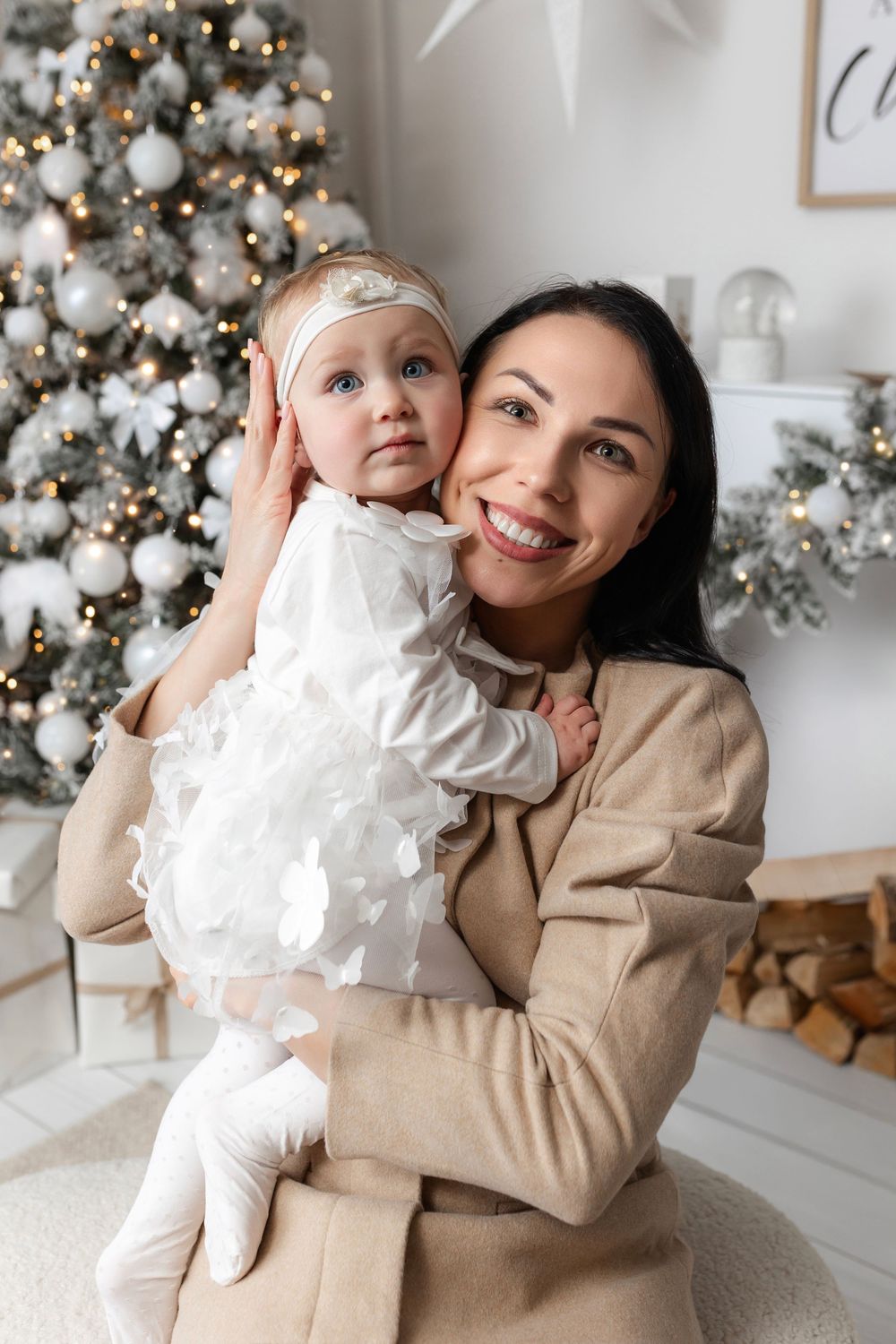Smiling mother holding baby girl in white dress near decorated Christmas tree with silver ornaments.