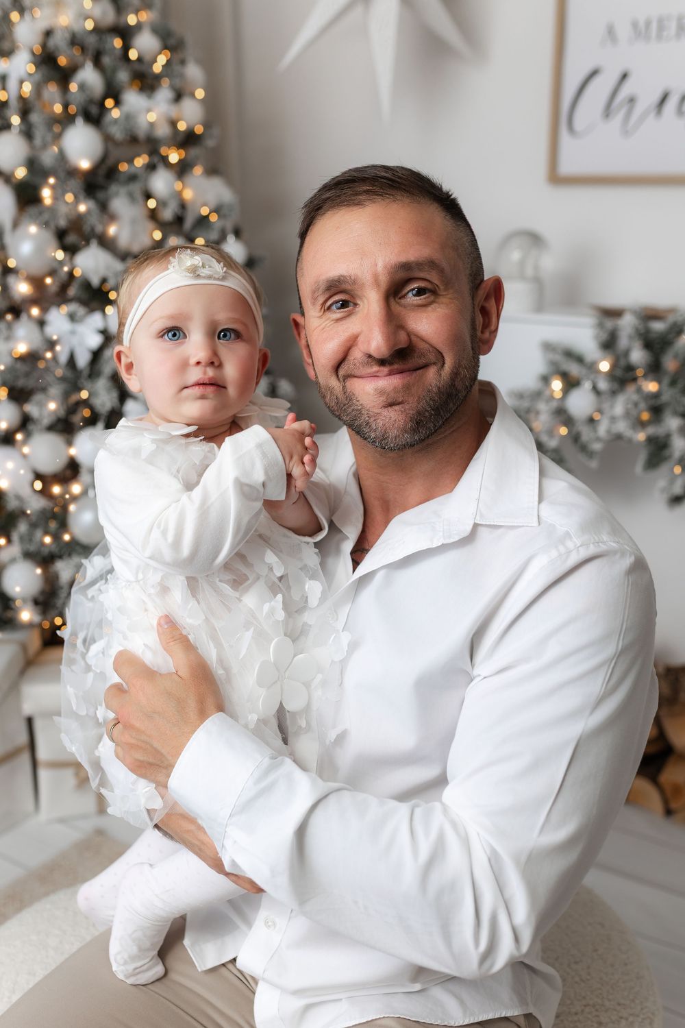 Father holding baby girl in white outfits posing together near decorated Christmas tree with warm lights.