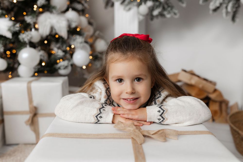 Young girl smiling behind a large Christmas gift box near a decorated white Christmas tree with warm lights.