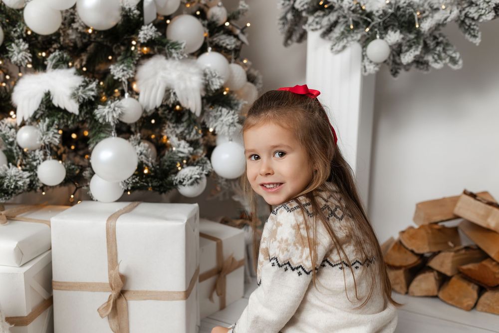 Young girl in festive sweater smiles beside wrapped Christmas gifts and decorated white tree with glowing lights.