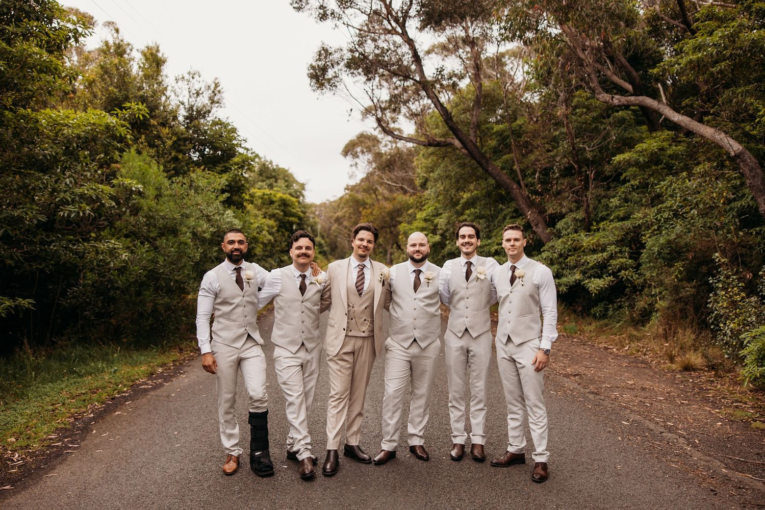 Groomsmen in light colored suits standing together on tree-lined path.