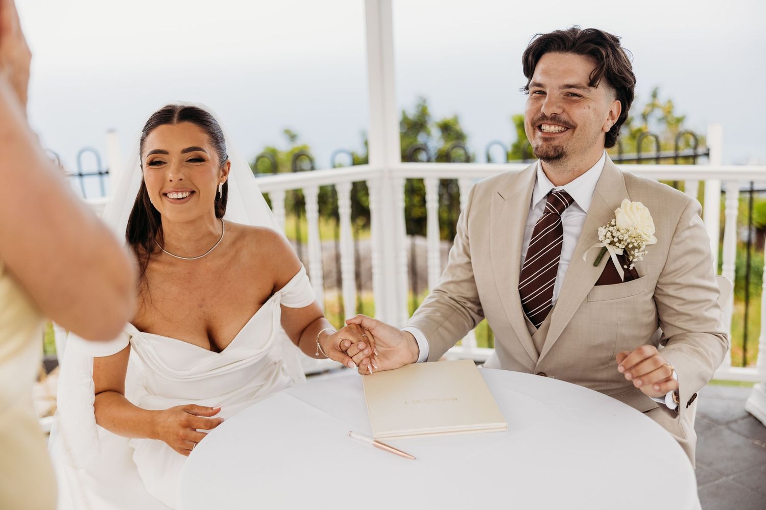A couple signs their marriage documents at a white table during their outdoor wedding ceremony.