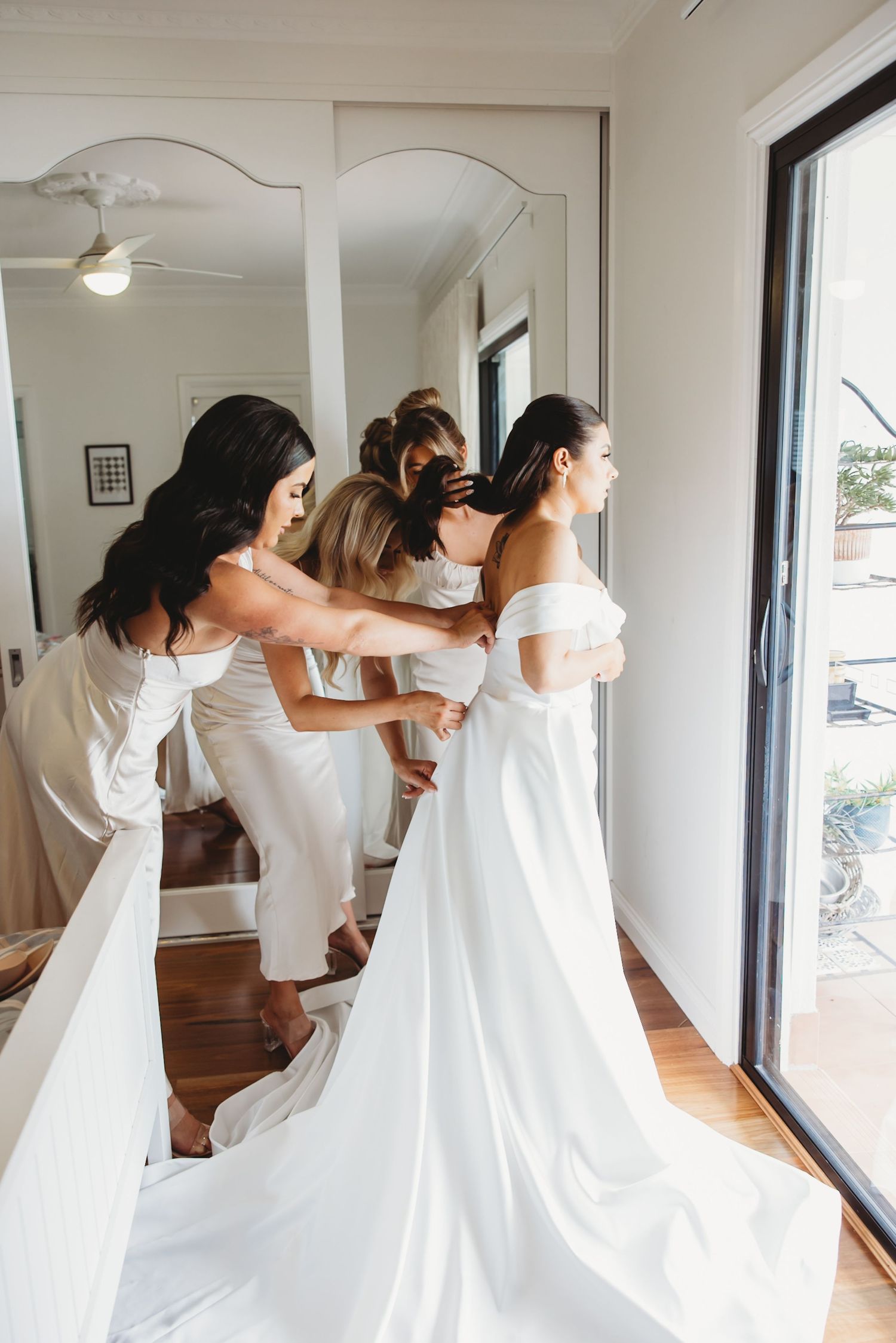 Bridesmaids in white dresses help adjust a wedding gown in a bright room with natural lighting.
