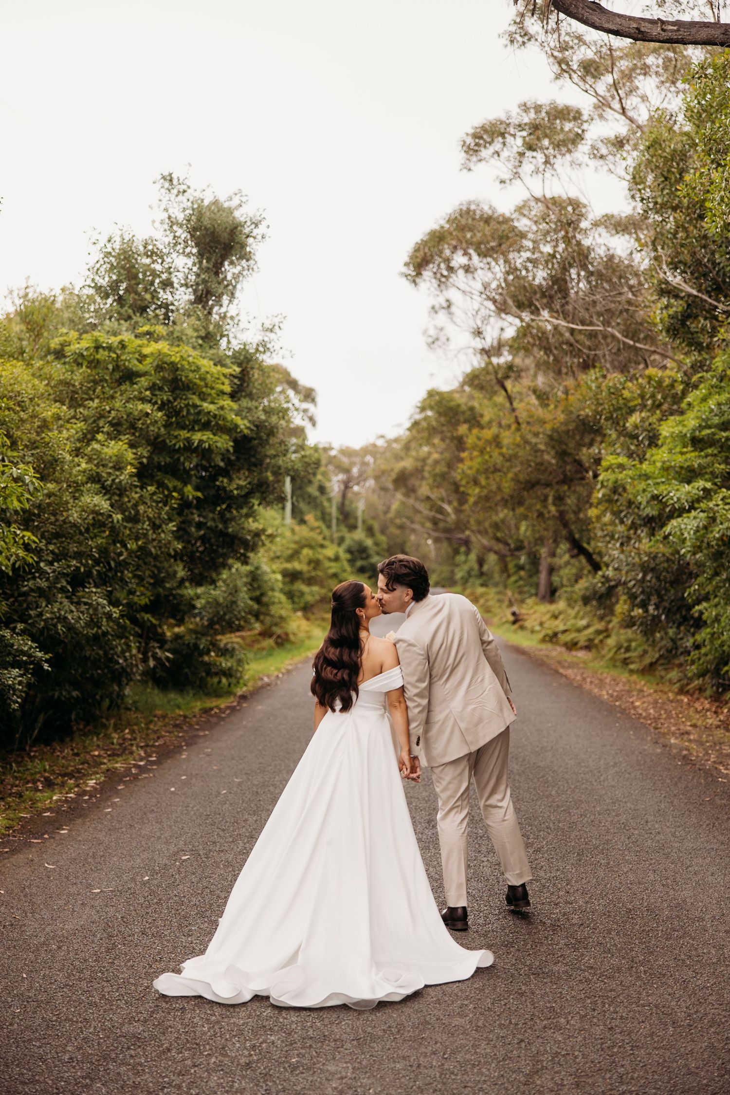 The newlyweds share a romantic moment on a tree-lined country road in formal wedding attire.