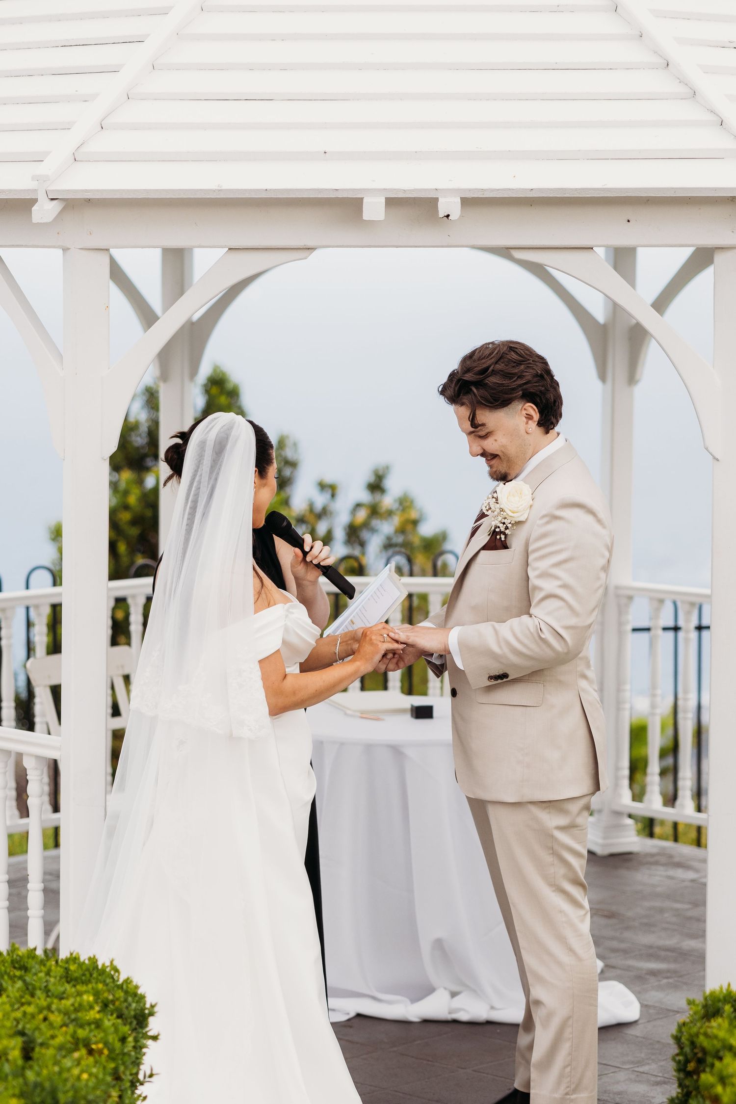 A wedding ceremony takes place under a white gazebo with floral decorations.