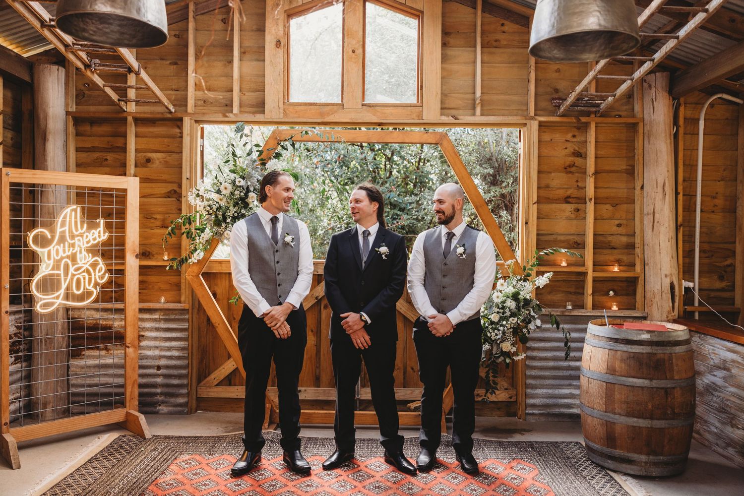 Three groomsmen stand in front of a floral arch inside a rustic wooden barn.