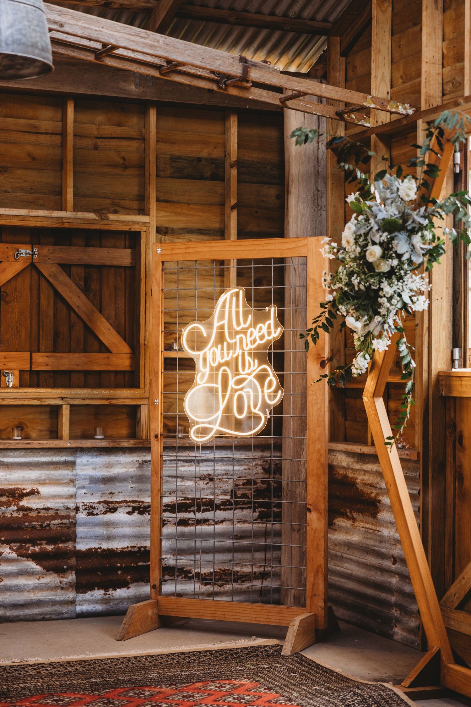 Illuminated 'All You Need is Love' sign with floral arrangements in rustic barn setting.