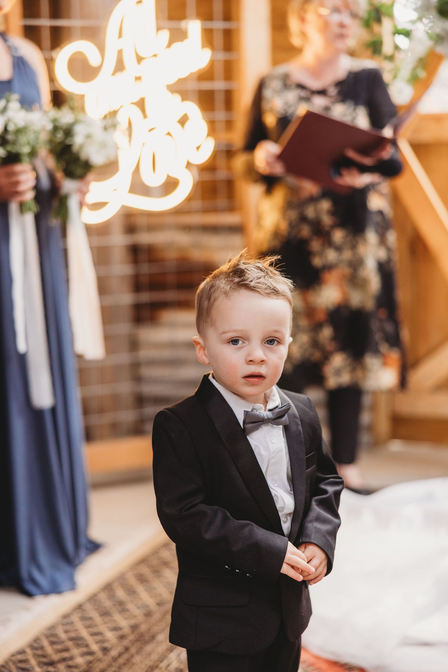 A young boy in a black tuxedo and bow tie stands during a formal wedding ceremony.