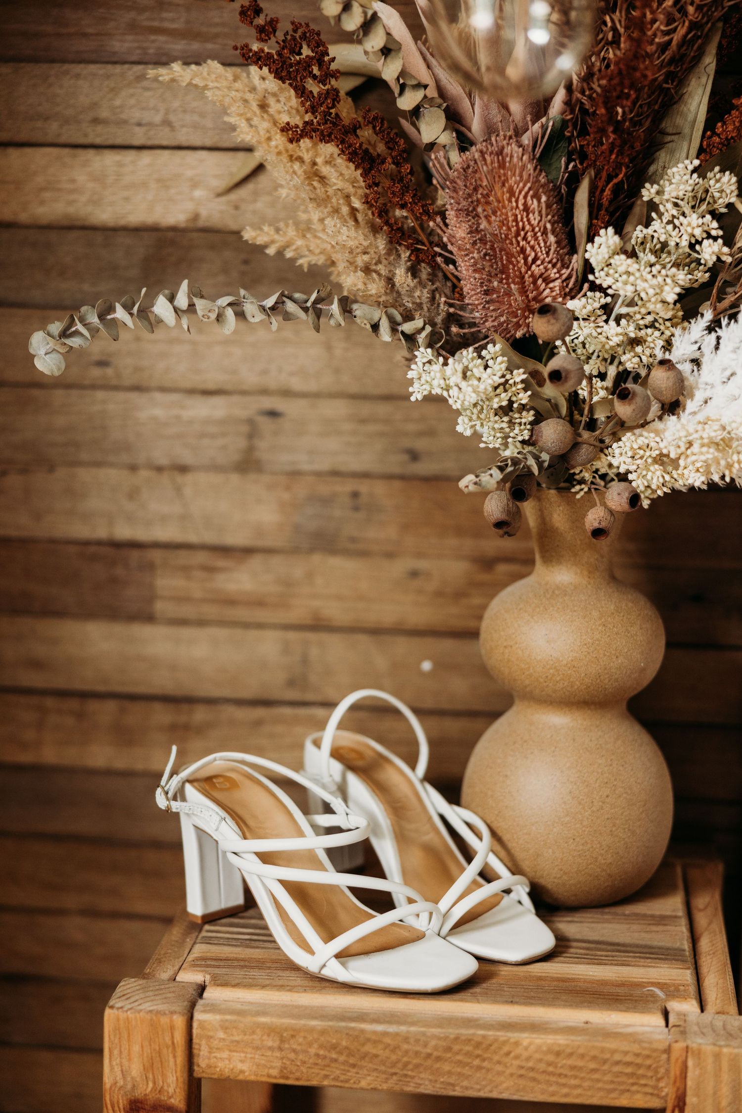 White bridal heels next to dried flower arrangement in beige vase on wooden surface.