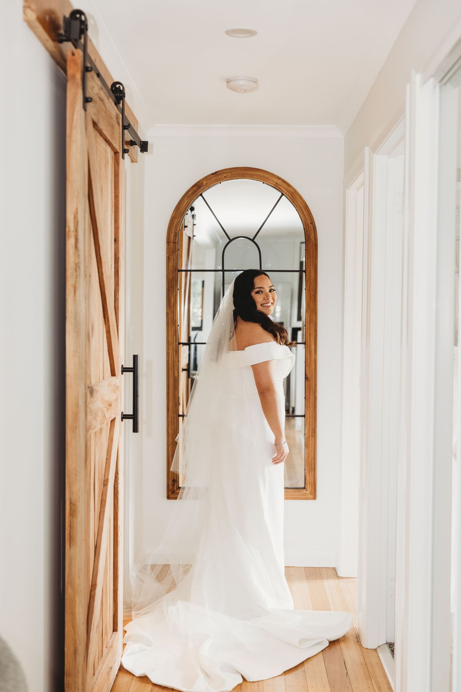 Bride in off-shoulder wedding gown poses in hallway with arched mirror and barn door.