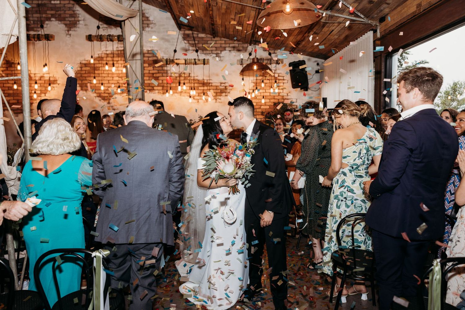 Wedding guests celebrate with confetti inside a rustic venue decorated with string lights.