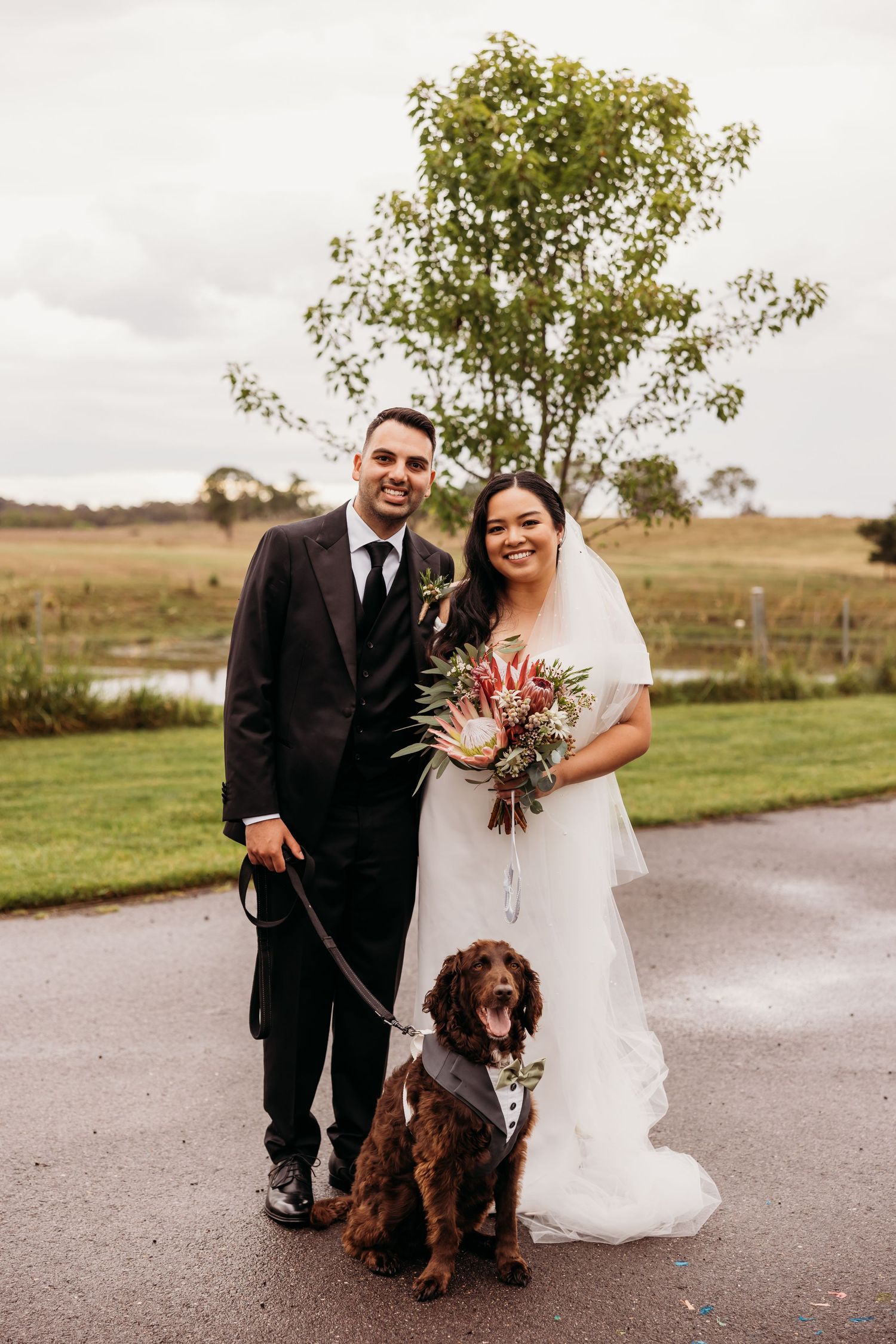 Newlyweds pose with their brown dog on a pathway with autumn landscape in background.