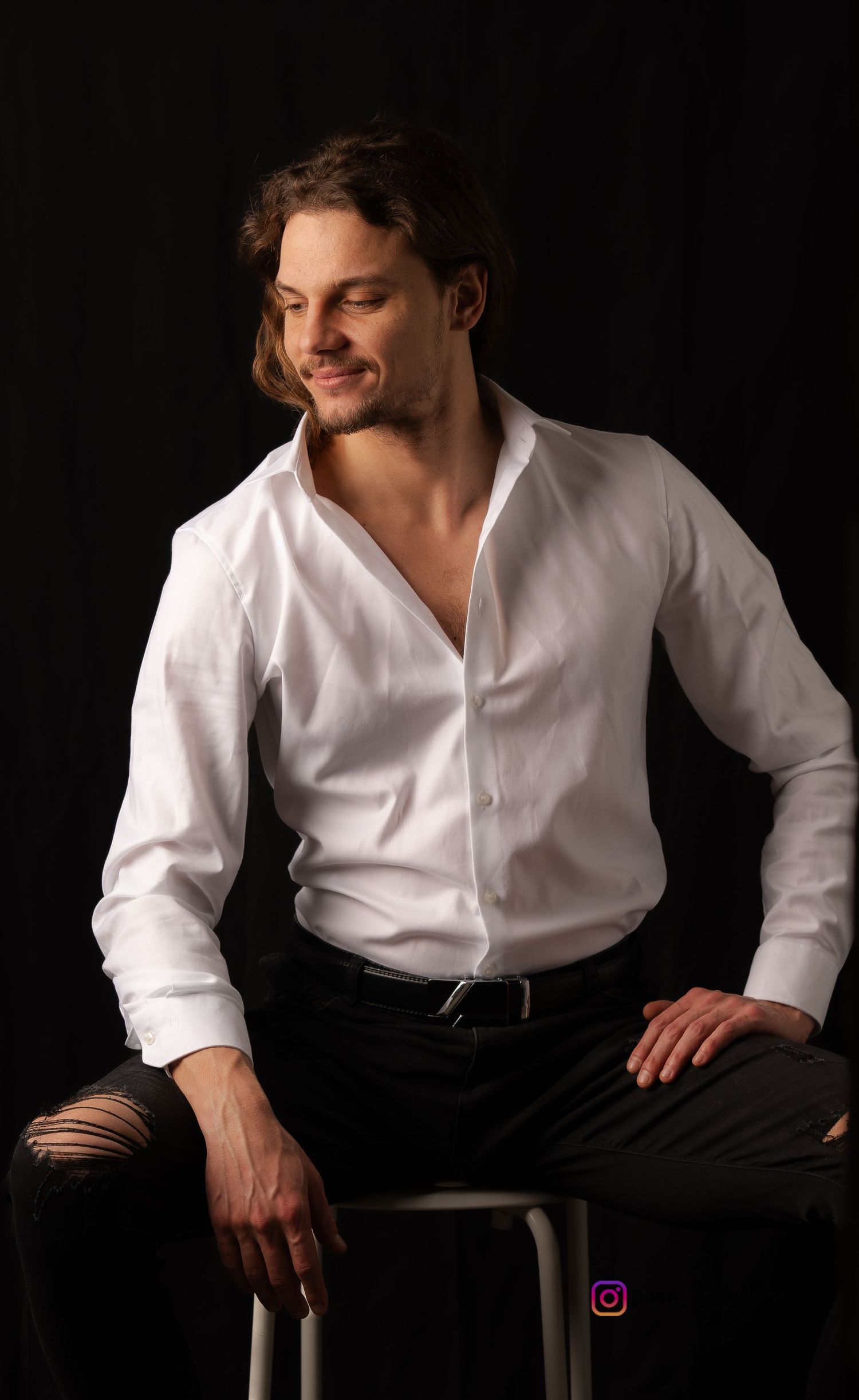 Dramatic studio portrait of person in white shirt and dark pants seated on stool.