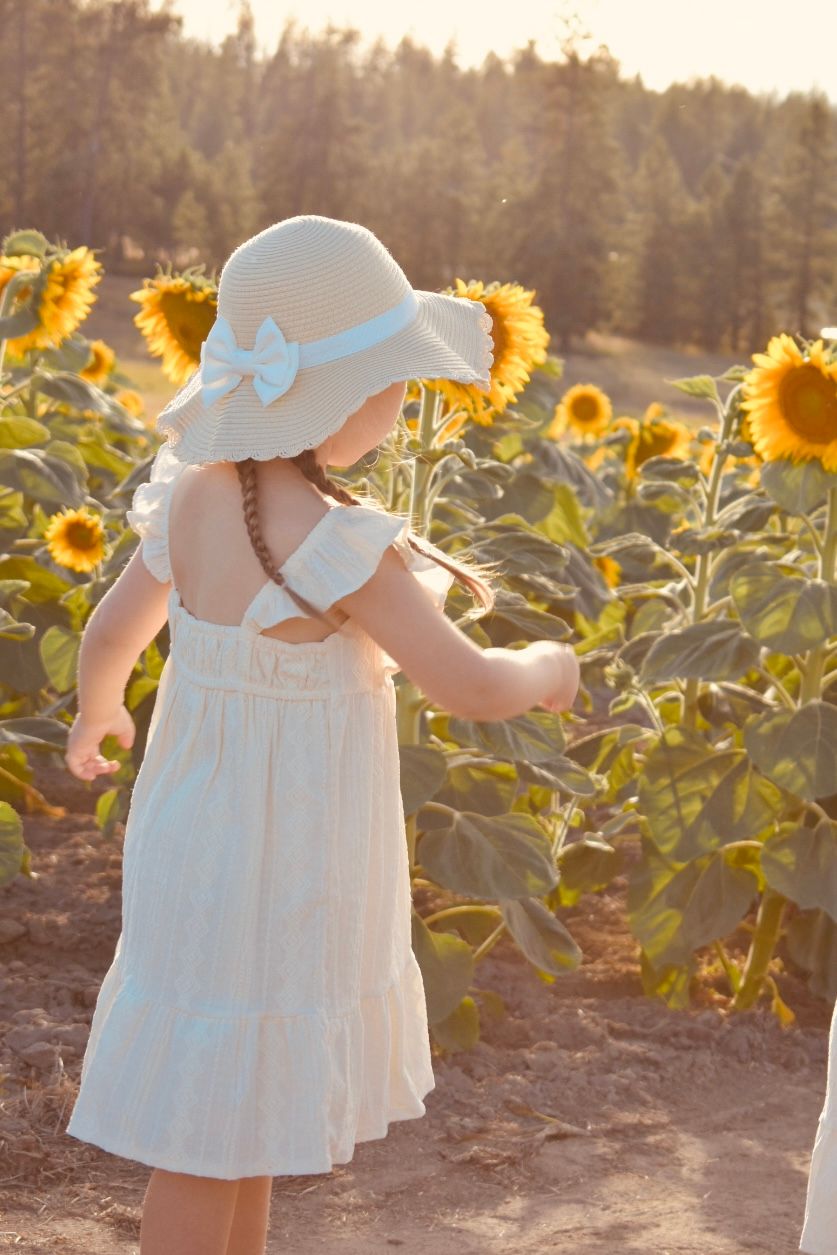 Sun-drenched figure in white dress and floppy hat exploring vibrant sunflower field at golden hour.
