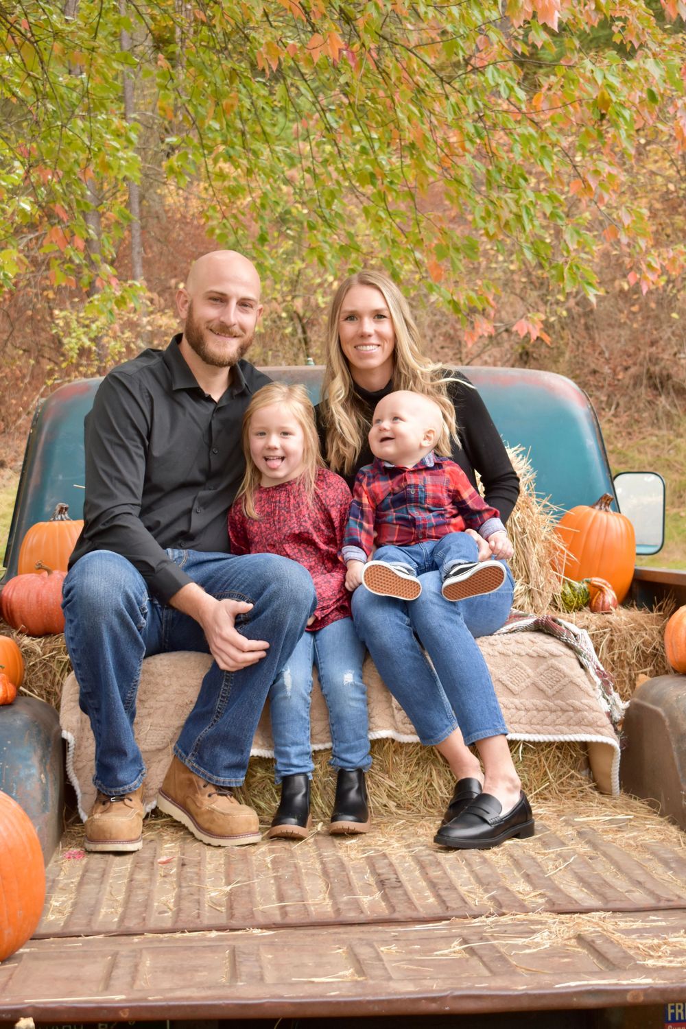 A family sits together on the bed of a vintage truck surrounded by pumpkins and hay bales.