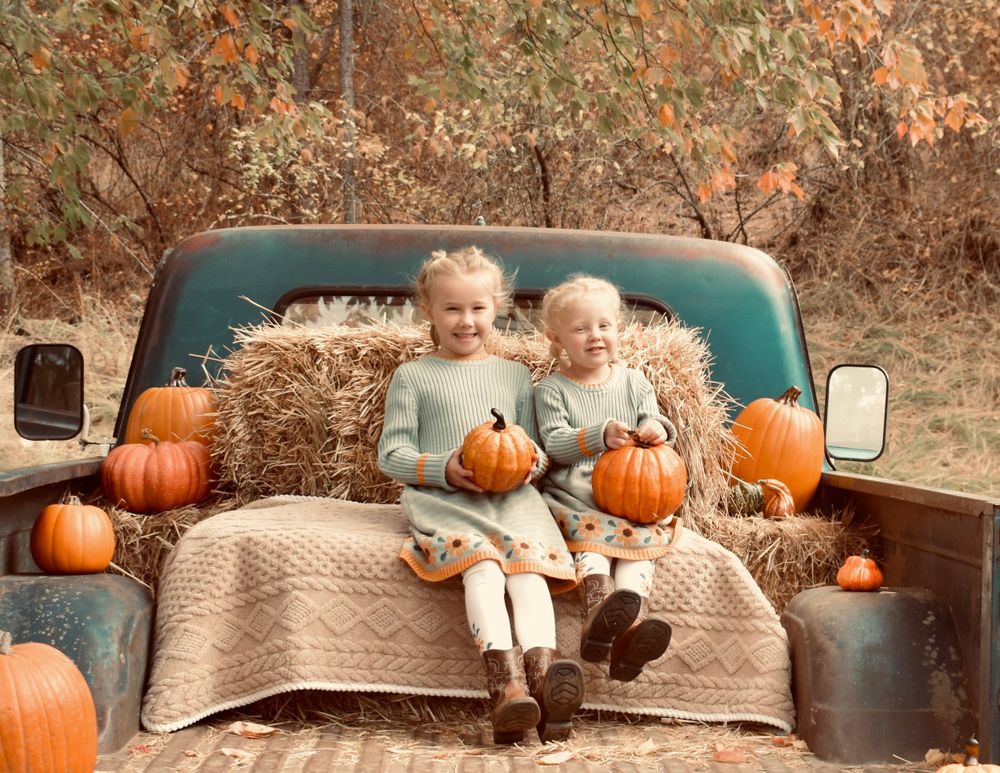 Autumn scene with children sitting on vintage truck bed surrounded by pumpkins and hay bales.