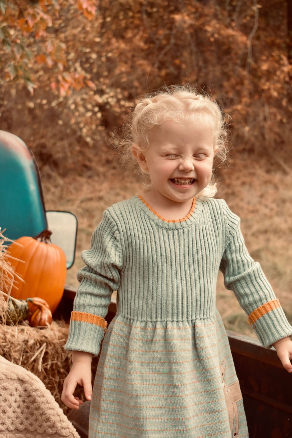 A joyful child wearing a sage green sweater dress poses near autumn decorations and hay bales.