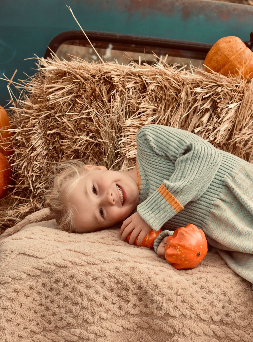 A smiling child lying on hay bales next to orange pumpkins in an autumn setting.