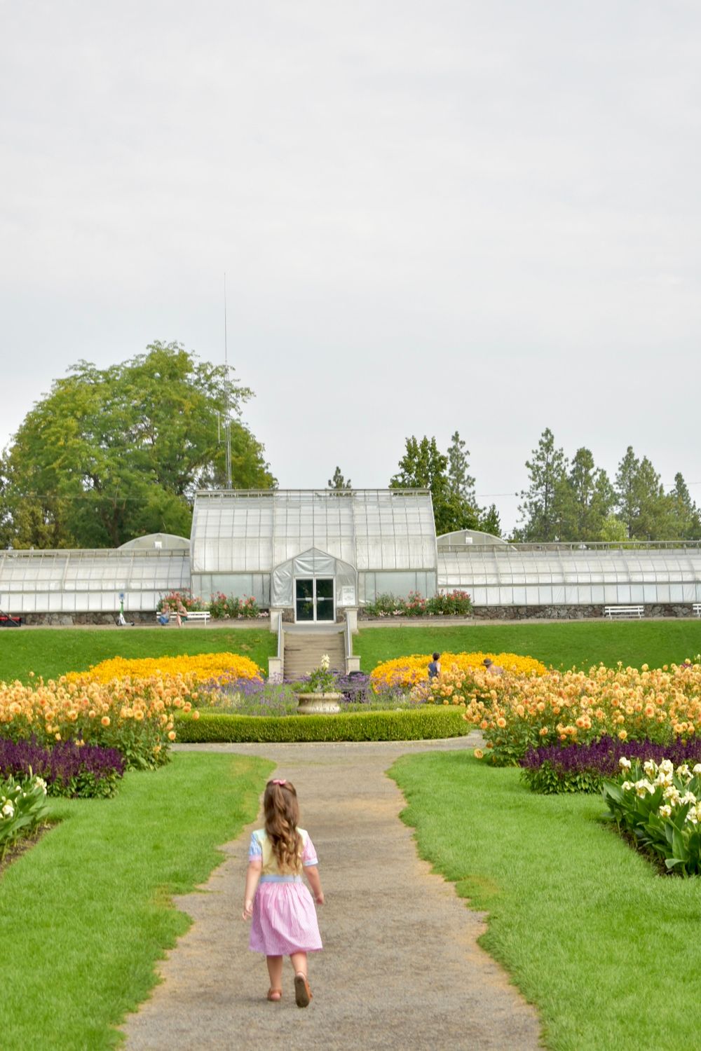 Young child walks along garden path toward greenhouse with vibrant tulip beds and manicured lawns at botanical garden.