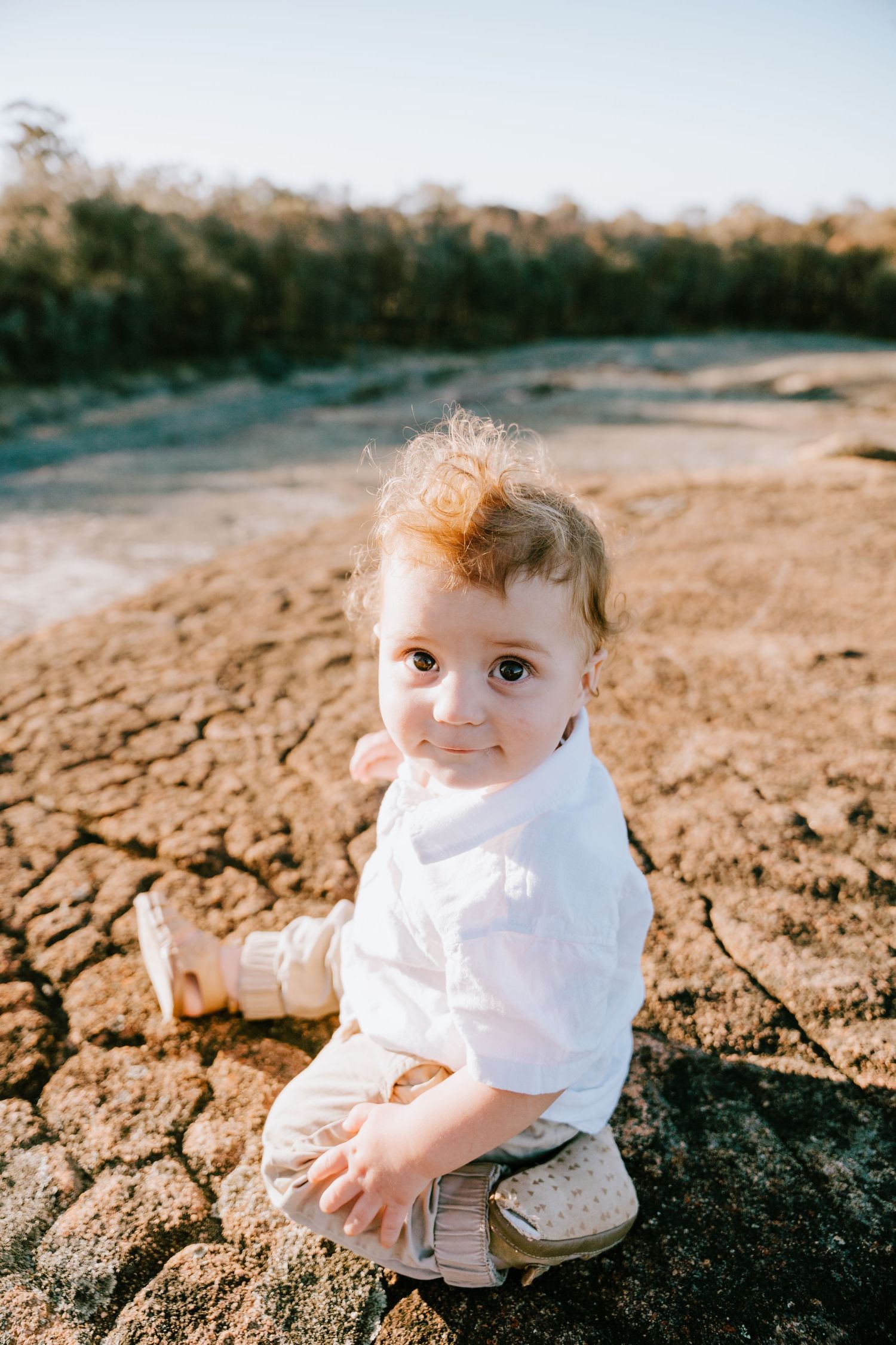 Baby in white outfit sitting on cracked desert ground during golden hour photography session.