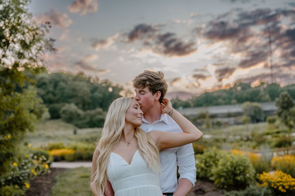 Couple sharing a romantic kiss at sunset in a lush garden, woman in white dress with long blonde hair.