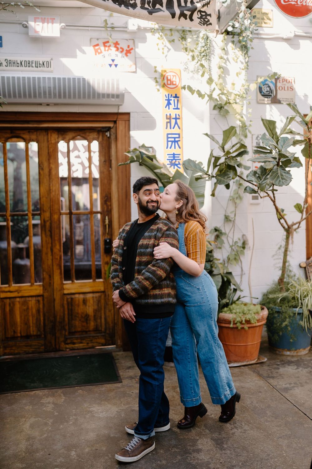 A couple embraces and smiles outside a charming shop with vintage signs and lush potted plants.