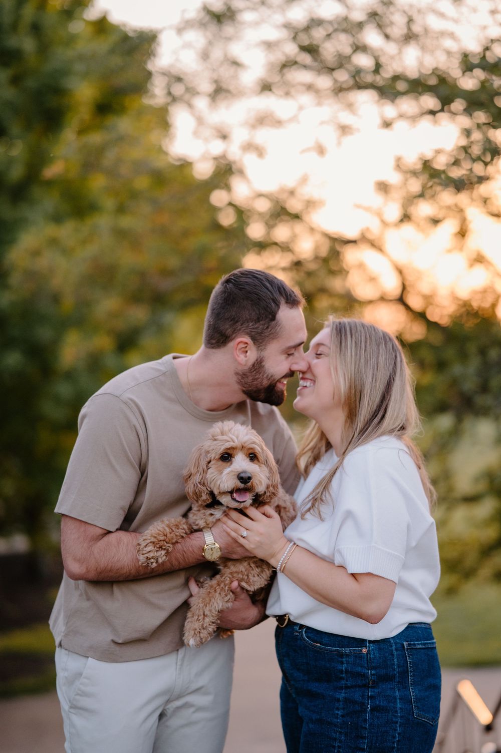 Couple sharing a kiss while holding an adorable golden doodle puppy outdoors at sunset.